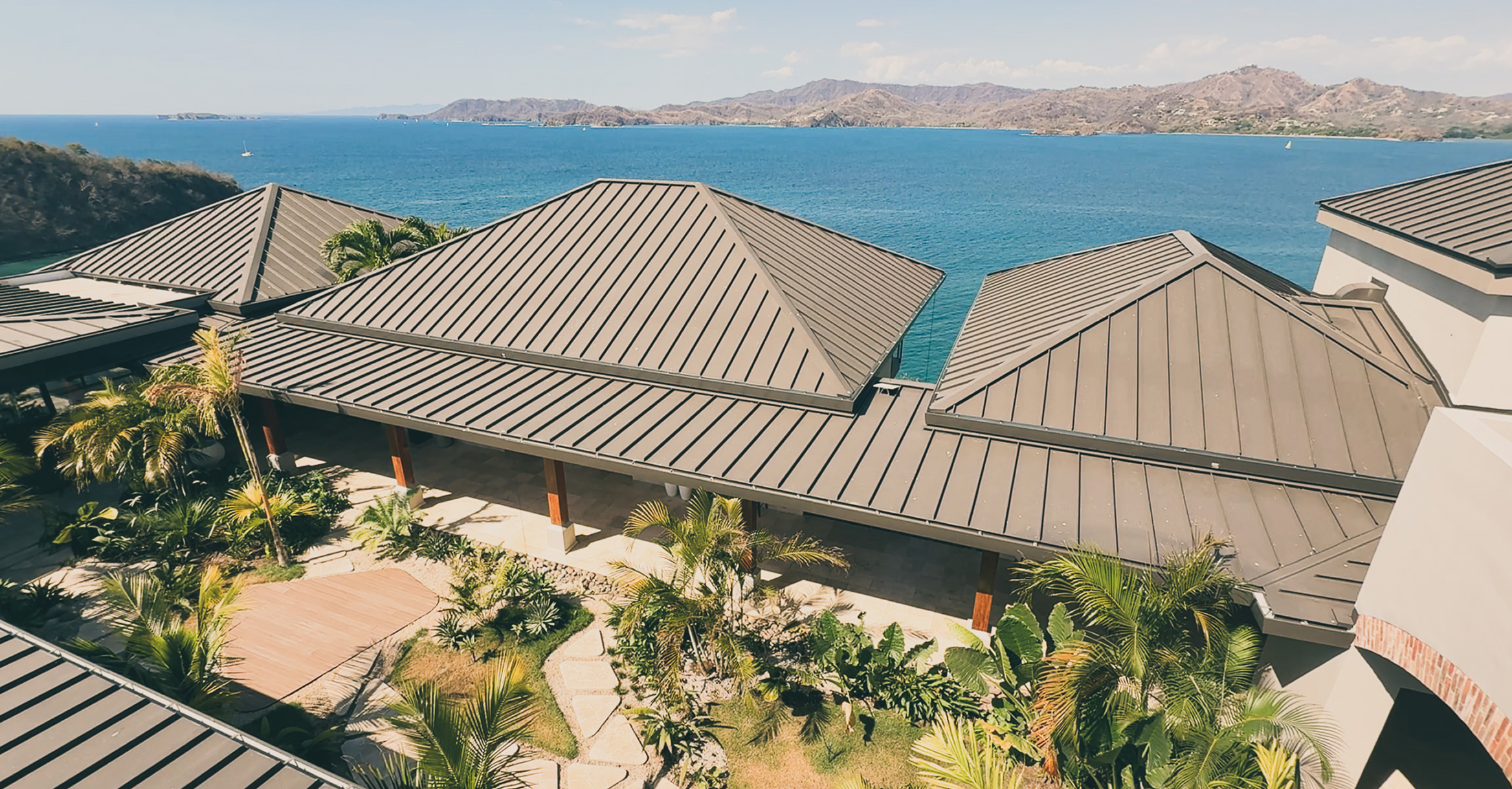 The villa's hipped metal rooftops and central courtyard garden, with the bay beyond.